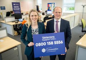 Woman and man hold a rectangular sign reading 'Veterans Support Line NI' with the telephone number 0800 158 5556 below. They are standing in front of a large desk with a number of people operating telephones