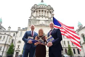 US Consul General, Belfast Lord Mayor and Communities Minister in front of City Hall with USA flag flying in background and all three holding books