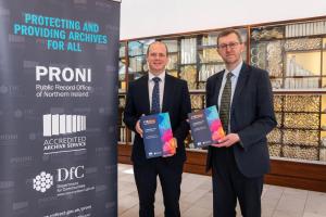 Two men wearing suits stand beside a banner which reads 'PRONI: Protecting and providing archives for NI'