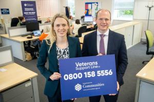 Woman and man hold a rectangular sign reading 'Veterans Support Line NI' with the telephone number 0800 158 5556 below. They are standing in front of a large desk with a number of people operating telephones